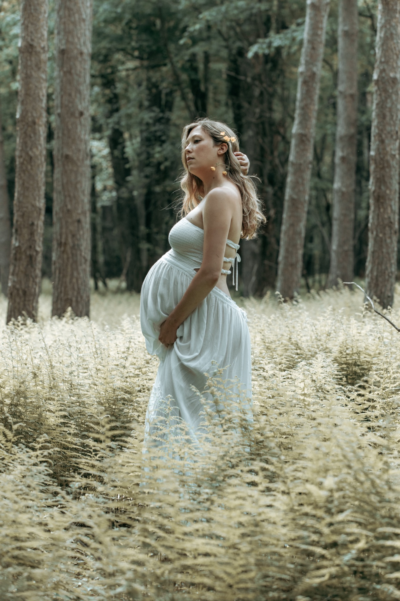 Expectant mother in a white dress standing among woodland ferns with small flowers in her hair