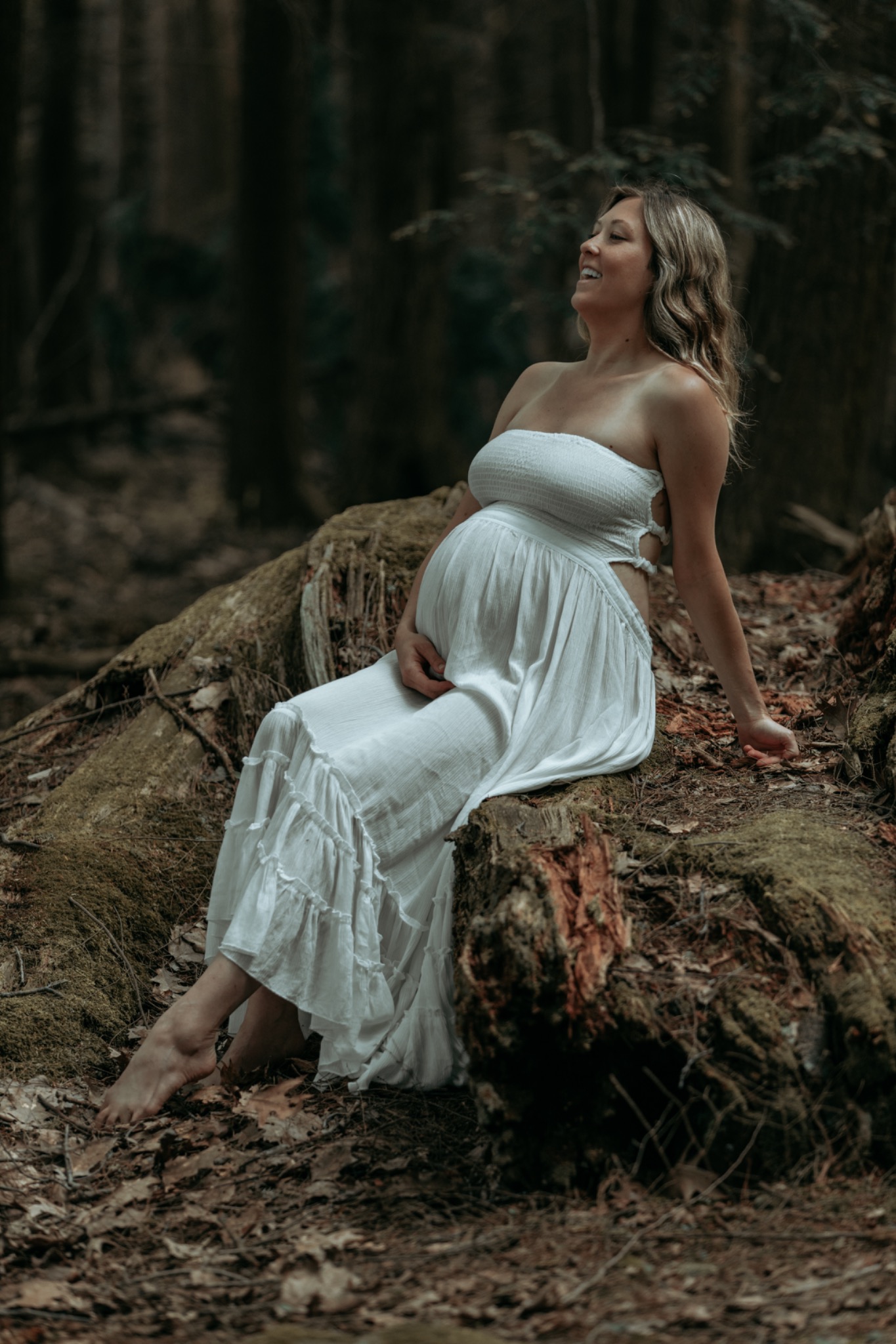 Expectant mother in a white dress sitting on a fallen tree in the woods