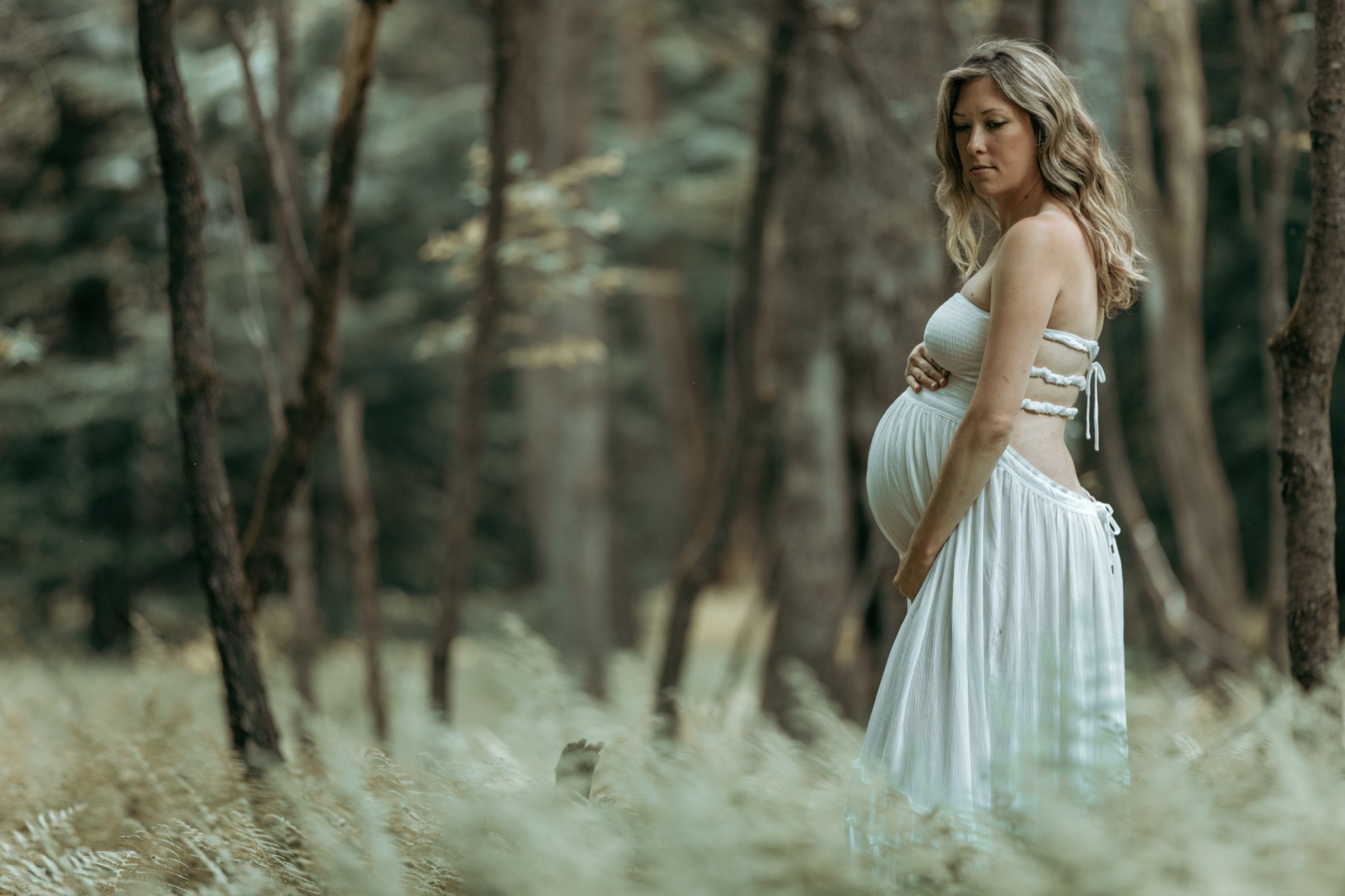 Expectant mother in a white dress standing among trees and soft woodland grasses