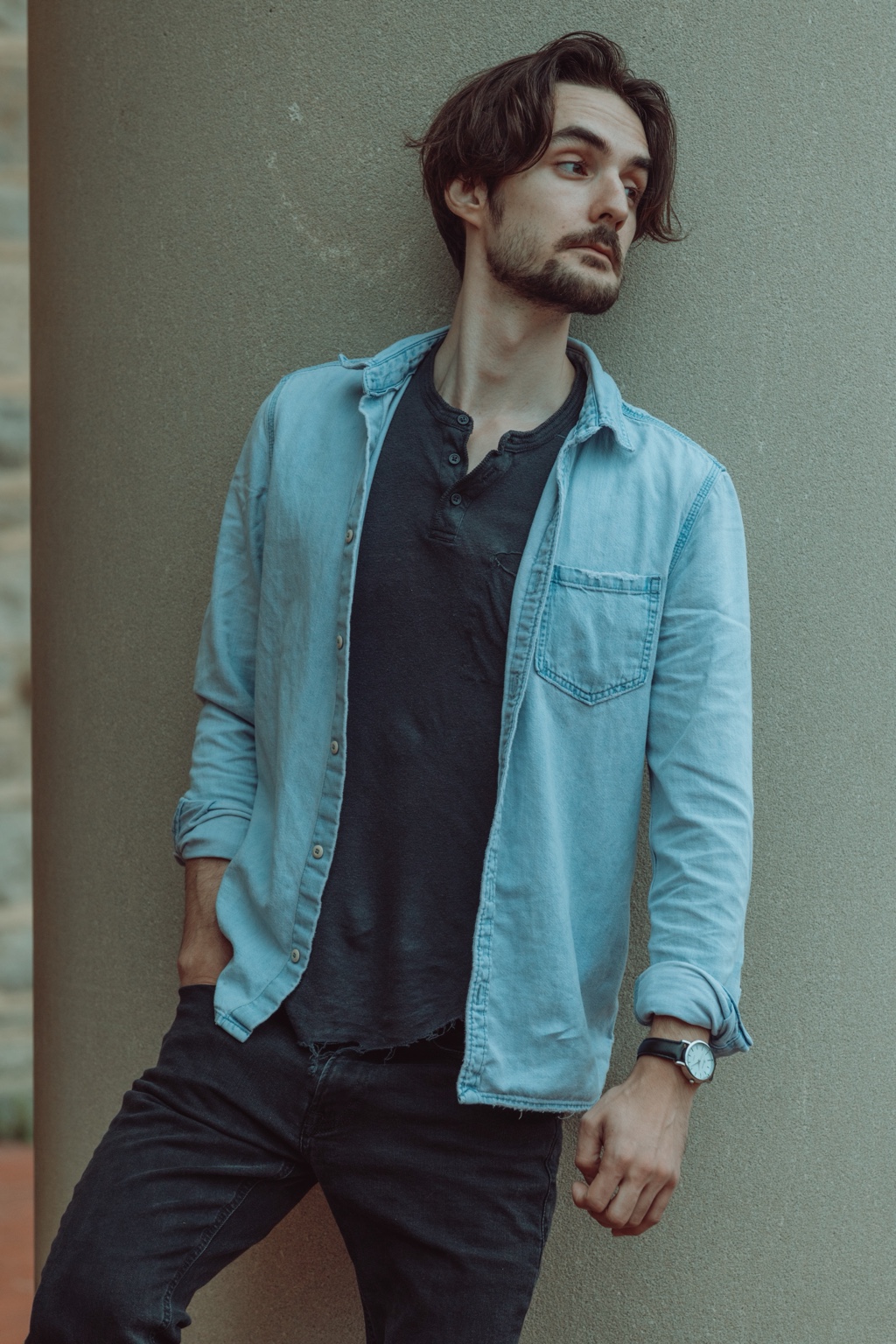 Man in a denim shirt standing against a textured wall and looking to the side